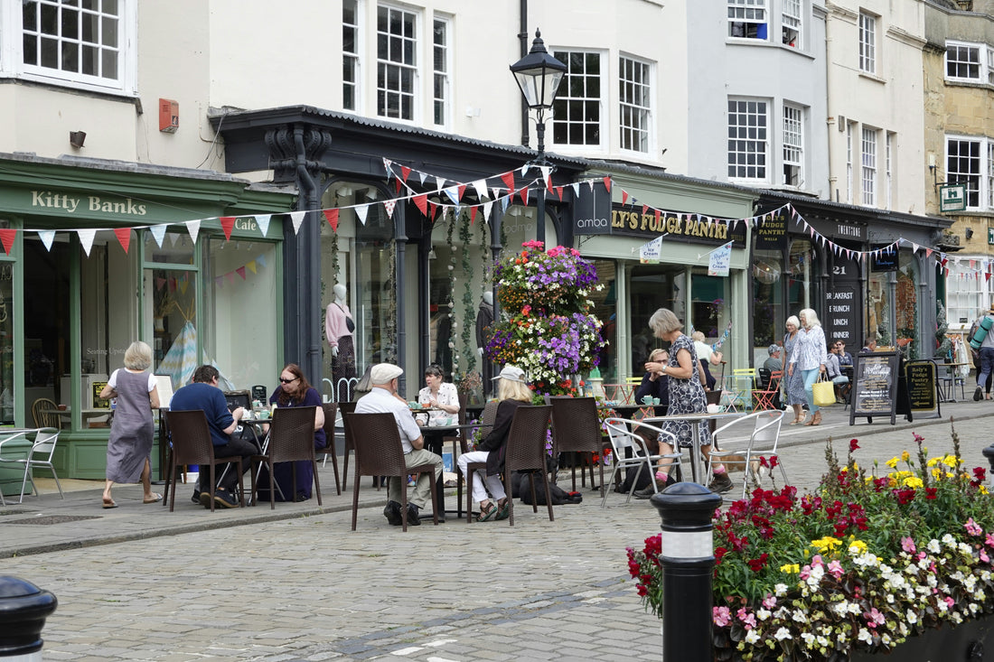 People dining at outdoor cafe tables along a charming cobblestone street lined with small shops, colorful flower displays, and festive bunting on a summer day.