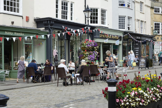 People dining at outdoor cafe tables along a charming cobblestone street lined with small shops, colorful flower displays, and festive bunting on a summer day.
